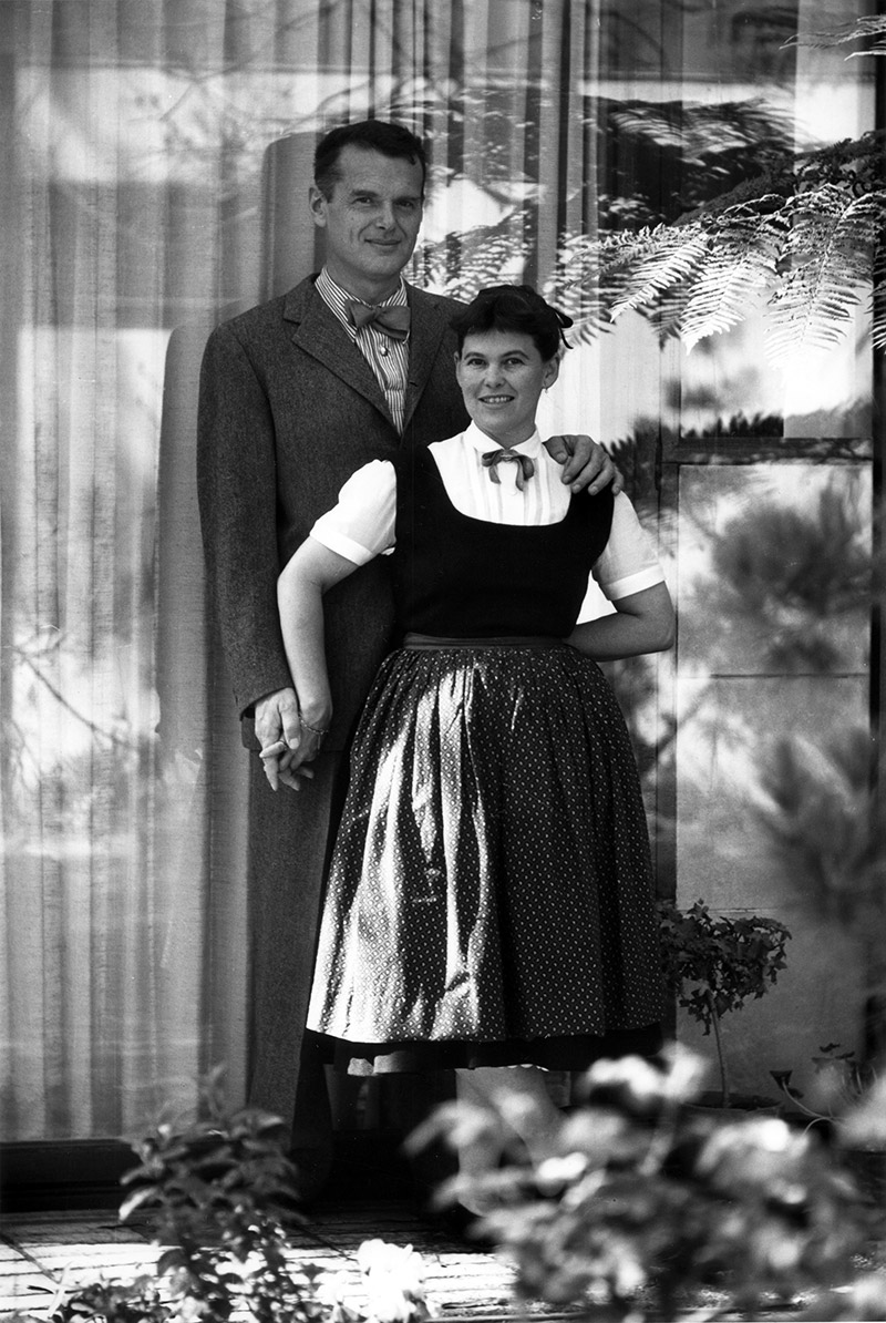 Black and white portrait of Charles & Ray Eames posed in front of the Eames House; they are holding hands